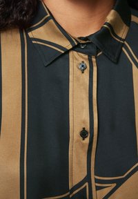 Skjorte med knapper med et sort og brunligt stribet design, med en spredt krave og sorte knapper. Stoffet virker glat og let struktureret.