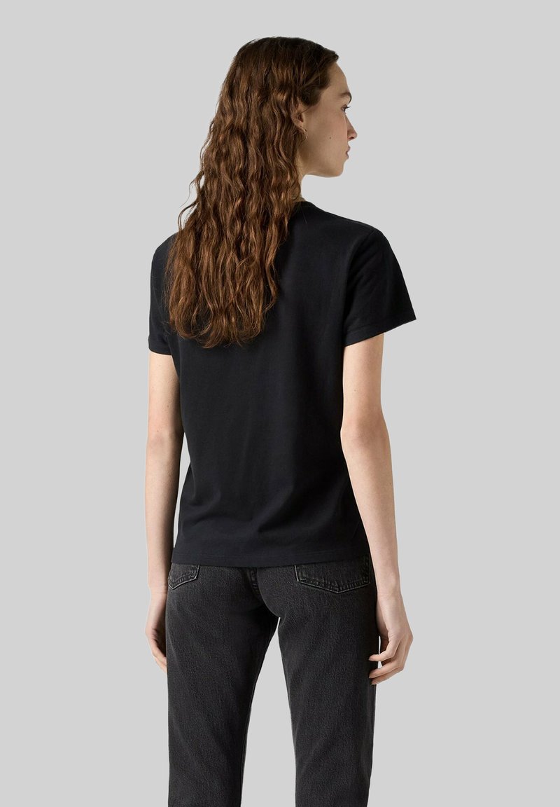 Frau mit langen, welligen Haaren, die ein einfaches schwarzes T-Shirt und dunkle Jeans trägt, steht mit dem Rücken zur Kamera vor einem grauen Hintergrund.