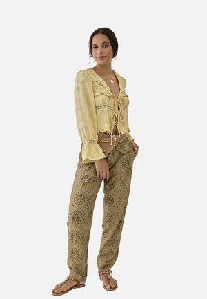 Mujer de pie con las manos en los bolsillos de pantalones estampados, lleva una blusa amarilla con cordones, un bolso marrón de cinturón y sandalias.