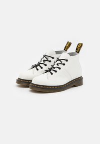 Botas de tobillo de cuero blanco con cordones negros, lazos de marca amarillos, costuras naranjas y una suela de goma texturizada.