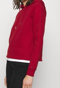 Persona che indossa una felpa rossa con cappuccio con "Tommy Hilfiger" ricamato sul manica e pantaloni neri, in piedi davanti a uno sfondo neutro.