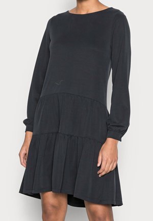 Femme portant une robe noire à manches longues, à volants, longueur genou, avec un col rond, sur un fond blanc uni.