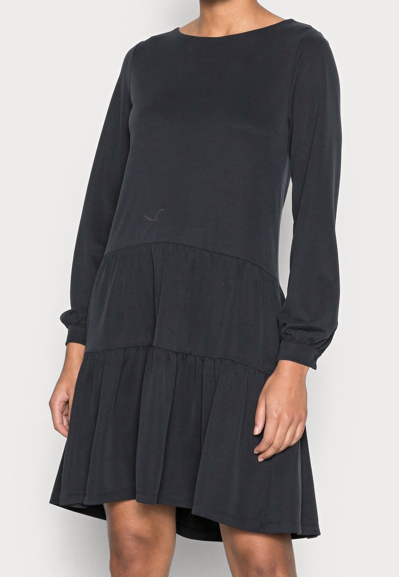 Femme portant une robe noire à manches longues, à volants, longueur genou, avec un col rond, sur un fond blanc uni.