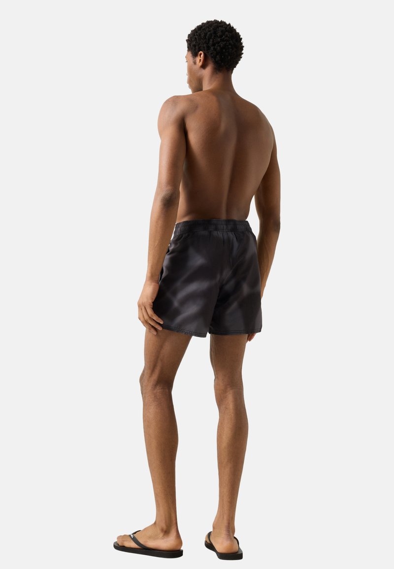 Homme debout pieds nus, de dos, portant un short de bain noir et des tongs noires sur un fond clair uni.