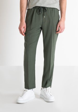 Mężczyzna ubrany w oliwkowozielone spodnie ze ściągaczem i białe trampki, stojący na tle jednolitego białego tła.