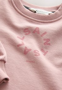 Lähikuva vaaleanpunaisesta collegeneuleesta, jossa on resoroitu kaulus ja hihansuut. Rintamuksessa näkyy pyöreä "AllSaints London" -logo haalistunein punaisin kirjaimin kankaassa.