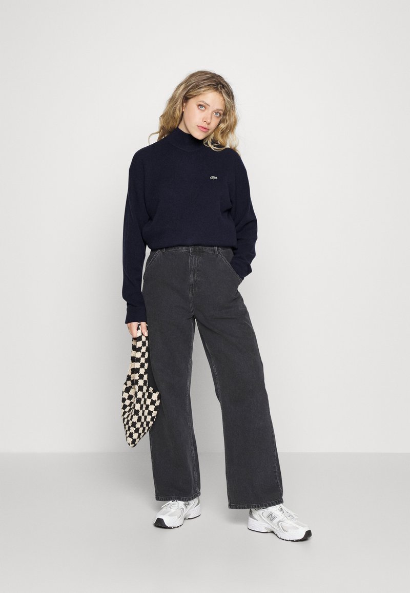 Pull à col roulé navy, jeans noirs à jambes larges et baskets blanches. Sac à carreaux à la main, avec une texture douce et des couleurs contrastées.