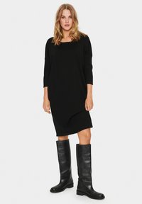 Vestito nero a lunghezza ginocchio con scollo quadrato e maniche a tre quarti, abbinato a stivali neri in pelle alti, creando un look minimalista.
