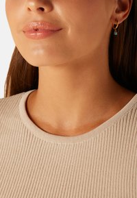 Gouden creool met een kleine turquoise hanger, gedragen met een lichtbeige ribgebreide top. Glad huid en een neutrale lipkleur zijn zichtbaar.