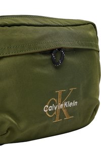 Bolsa de nailon verde con superficie texturizada, cierre de cremallera y detalle de cordón negro. Logo bordado de "Calvin Klein" en dorado y blanco.