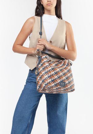 Donna che indossa un gilet beige e jeans blu tiene una borsa a tracolla intrecciata multicolore con motivo blu, marrone, bianco e tracolla in pelle.