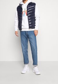 Man i en vit hoodie med färgglada bokstäver, marinblå puffer väst, blå uppvikta jeans, vita strumpor och vita sneakers står på en grå golv.