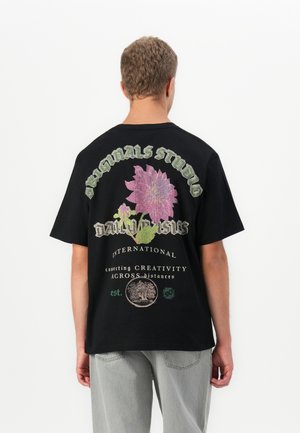 Ung mann iført svart oversized T-skjorte med stor rosa blomst og tekstgrafikk på ryggen, sammen med lysegrå jeans.