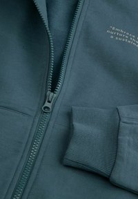 Zbliżenie na częściowo zapiętą na suwak miętową bluzę z prążkowanymi mankietami i częściowym białym napisem na piersi: "Embrace... nurture... a sustaina..."