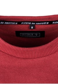 Red knitted sweater with a ribbed crew neckline, featuring a black label with white text and a size tag "M." Soft texture with a smooth finish.
