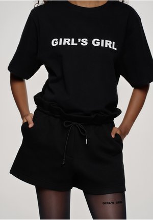 Osoba nosící černé oversized tričko a šortky s nápisem „GIRL'S GIRL“ na tričku a stehně, ruce v kapse.