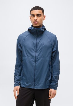 Giovane uomo che indossa una giacca a vento blu con cerniera e collo alto e pantaloni neri, in piedi contro uno sfondo chiaro e uniforme.