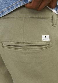 Olivově zelené kalhoty s hladkým povrchem, se zadní kapsou na zip a malou bílou etiketou s logem "JACK & JONES".