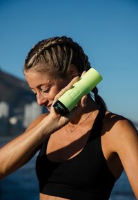 Femme en tenue de sport tenant un haut-parleur portable vert près de son visage en plein air, avec un ciel bleu et des bâtiments flous en arrière-plan.