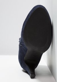 Bottine à talon haut en daim bleu marine à semelle noire, posée sur une surface blanche contre un mur blanc.