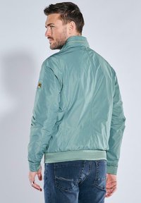 Hellgrüne Nylonjacke mit hohem Kragen, gerippten Manschetten und Saum. Mit Kontrastnähten und einem Logo-Patch am linken Ärmel.