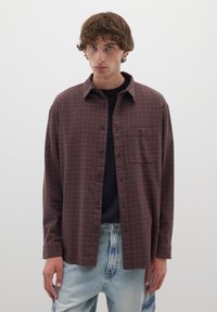 Camicia a scacchi marrone realizzata in tessuto morbido, con chiusura a bottoni, colletto e taschino sul petto. Indossata sopra una maglietta nera, abbinata a jeans chiari.