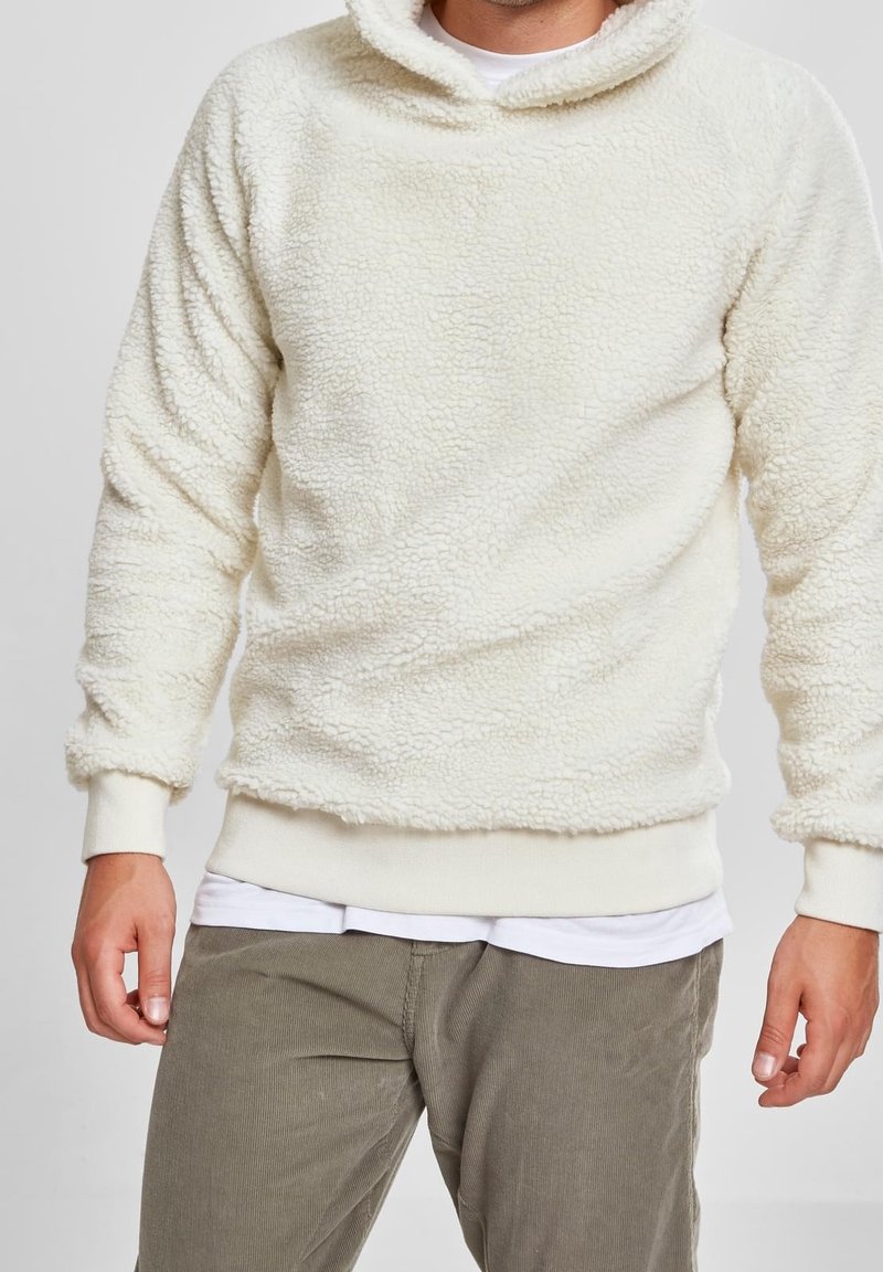 Cremefarvet fleece-pullover med en tekstureret, blød overflade, med hætte og ribbede manchetter. Blevet båret over en hvid skjorte og parret med olivengrønne bukser.