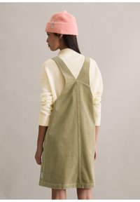 Sukienka ogrodniczka z prążkowanego sztruksu w kolorze oliwkowej zieleni o dopasowanym kroju, z szerokimi ramiączkami i dekoltem w kształcie litery V na plecach. Nosi się ją na kremowym swetrze, dopełniona różową czapką beanie.
