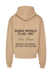 Beige színű kapucnis pulóver, nagy kapucnival, fekete szöveggel a hátán: "DUBAI WORLD CLUB, 1997," "Luxe Leisure," és "TISZTELD AZ EMBEREKET, NE CSAK A KULTÚRÁT."