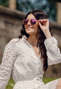 Bílá šaty s výšivkou, dlouhými rukávy a páskem v pase, doplněné světle růžovými slunečními brýlemi se zrcadlovými čočkami. Venkovní prostředí.