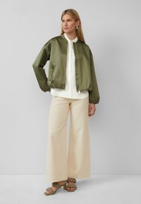 Veste bomber vert olive, tissu lisse, fermeture éclair, chemise blanche à boutons, pantalon large crème avec une texture subtile, et sandales olive.