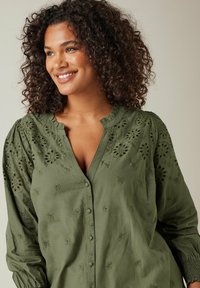 Olivegrüne Bluse mit floralem Stickerei, die eine durchgehend knöpfbare Vorderseite, einen Rüschensaum und gepuffte Ärmel mit elastischen Manschetten aufweist.