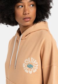 Hoodie aus weichem Pfirsichstoff, mit einem großen weißen kreisförmigen Logo und blauen Akzentgrafiken auf der Brust, mit Kordeln.
