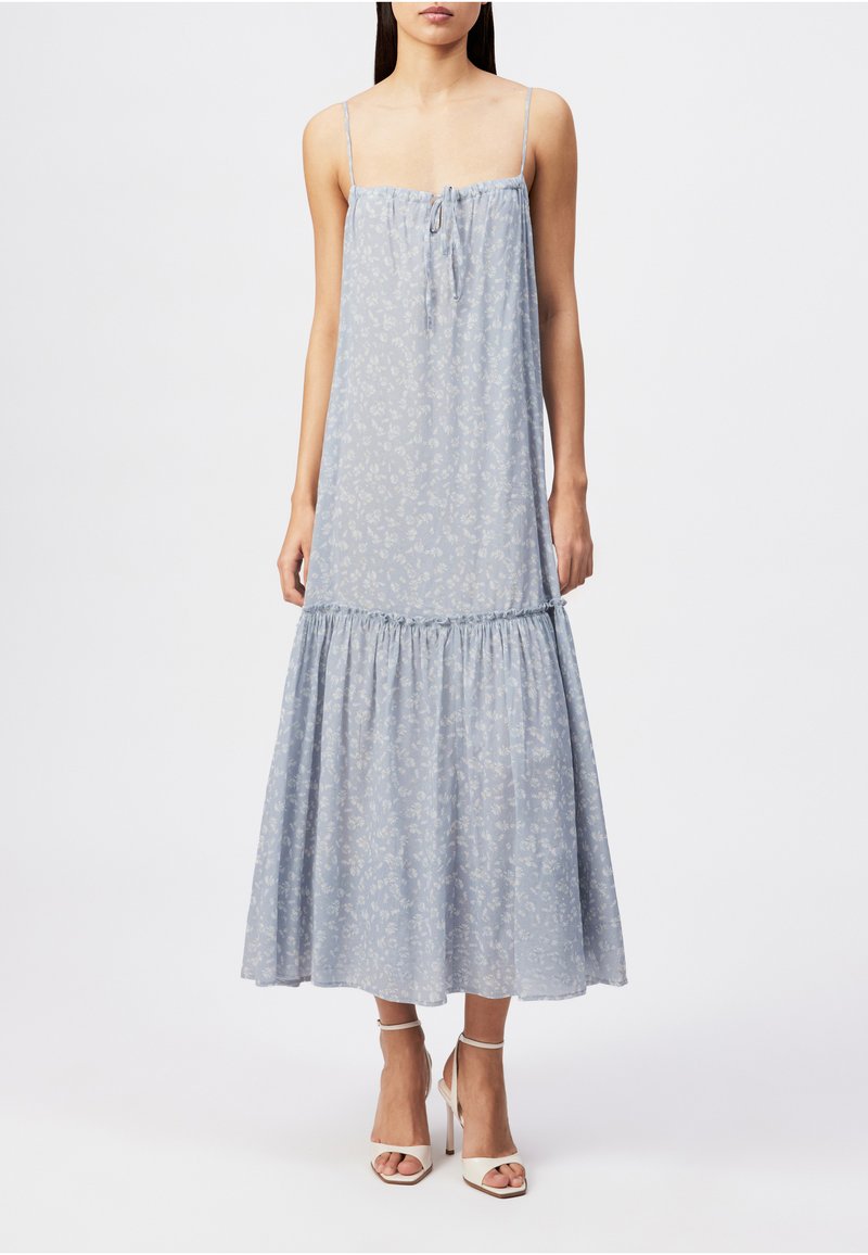 Femme portant une robe midi bleu clair à bretelles fines avec un motif floral, une jupe à volants froncés et des sandales blanches à talons hauts.