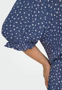 Zila ziedu kleita ar savilktām pūkainām piedurknēm. Balti līdzīgi margrietiņām raksti visā apģērbā, teksturēta auduma un viļņoti piedurkņu maliņas.