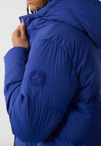 Blauwe pufferjack met een ritskap, met ribgebreide manchetten en een ronde patchlogo op de linkerarm. Glad, glanzend materiaal met een lichte vulling.