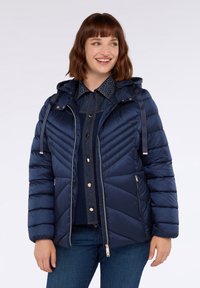 Donna sorridente che indossa un giubbotto imbottito in tessuto trapuntato blu navy con cappuccio, zip a metà, sopra una camicia in jeans con bottoni e jeans blu.