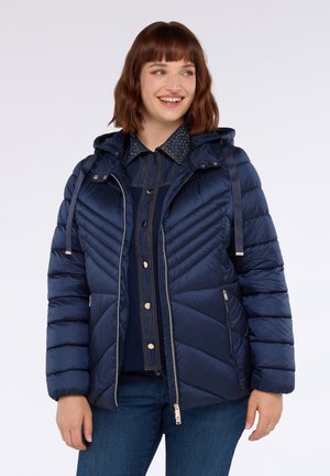 Glunderende vrouw in een marineblauwe gewatteerde pufferjack met capuchon, tot halverwege dichtgeritst, over een denim overhemd met knopen en een spijkerbroek.