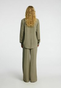 Olijfgroene, loszittende blouse met lange mouwen, gecombineerd met een broek met wijde pijpen. Glad, textuurgedragen stof; gebogen onderrand op de top.