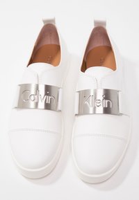 Vita slips-on skor med en slät läderöverdel. Har ett silverfärgat metallband med "Calvin Klein" utskuren bokstäver överst.