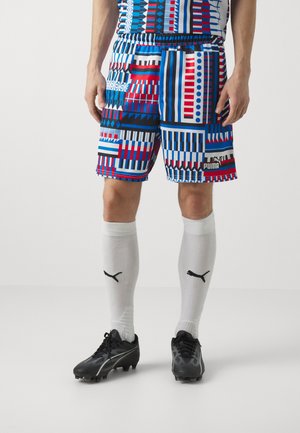 Shorts de sport Puma avec un motif géométrique multicolore en bleu, rouge et blanc. Dotés d'une taille élastique et d'un logo. Associés à des chaussettes blanches et des crampons noirs.