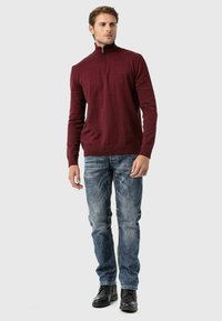 Burgunderfarbener Pullover mit Reißverschluss, aus weichem Material, kombiniert mit hellblauen Jeans und schwarzen Stiefeln. Hat ein tailliertes Design mit gerippten Bündchen.
