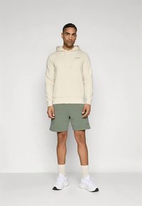 Μπεζ φούτερ με κουκούλα και μπροστινή τσέπη, σε συνδυασμό με χακί σορτς. Λευκά αθλητικά παπούτσια και ανοιχτόχρωμες κάλτσες μέσης συμπληρώνουν την εμφάνιση.