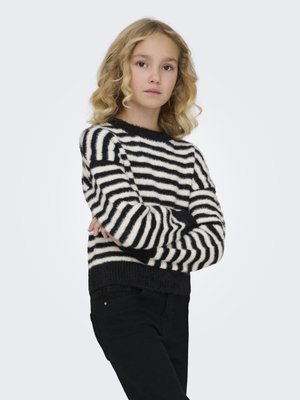 Fille aux cheveux blonds bouclés, debout, bras croisés, portant un pull duveteux rayé noir et blanc et un pantalon noir sur un fond uni.