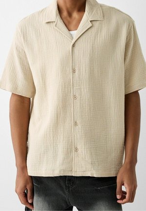 Homme portant une chemise beige à manches courtes texturée et boutonnée par-dessus un top blanc avec un jean noir délavé, debout devant un fond neutre.