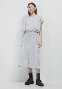 Donna in piedi che indossa un top cinch grigio chiaro e una gonna midi con stivali combat neri con lacci, su uno sfondo bianco semplice.