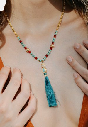 Gold chain necklace featuring turquoise and red beads, with a gold pendant and a teal tassel, resting on bare skin.