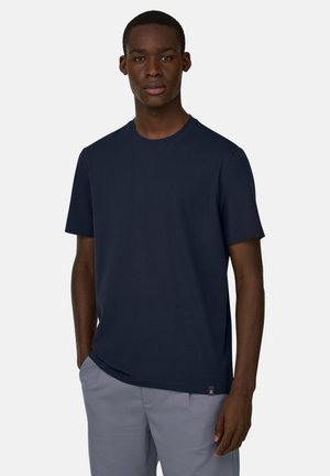 Ung mand iført en ensfarvet mørkeblå kortærmet t-shirt og lysegrå bukser, stående med den ene hånd i lommen mod en hvid baggrund.