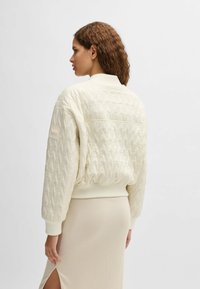Cream quilted bomber jacket with ribbed cuffs and hem, featuring a subtle logo patch on the sleeve and a relaxed fit.