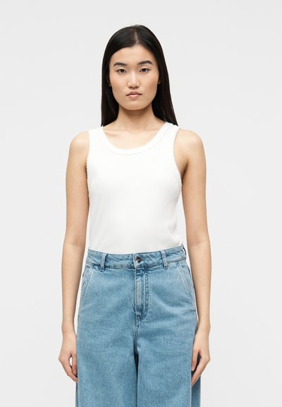 Jeune femme aux longs cheveux bruns portant un haut blanc sans manches et un jean en denim bleu clair taille haute, debout devant un fond uni.
